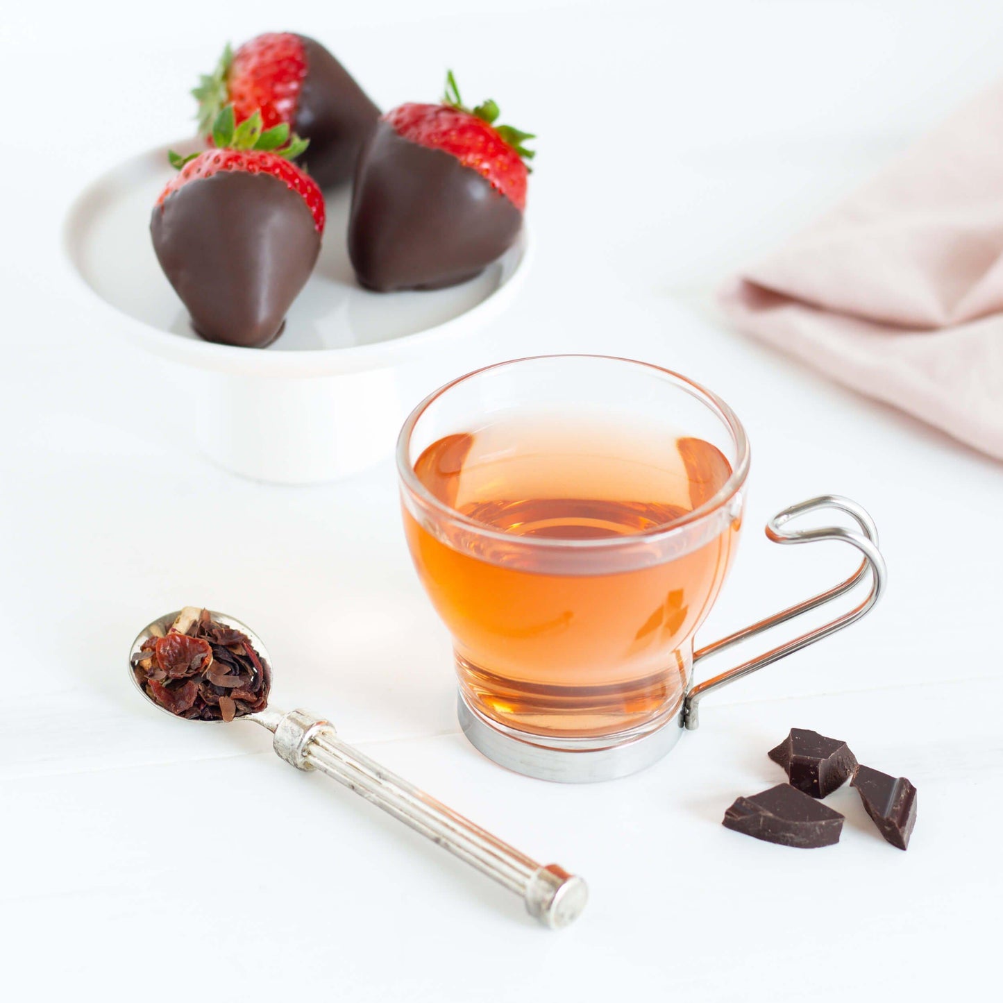 Chocolate Strawberry Organic Herbal Tea shown as brewed tea in a glass mug with a metal handle. A dish with three chocolate-covered strawberries is in the background. A spoon of tea leaves and few pieces of chocolate are in the foreground