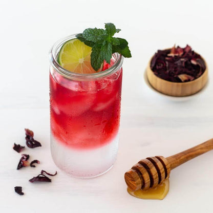 Hibiscus Organic Herbal Tea shown as iced tea with a slice of lime and mint leaves. Loose hibiscus tea leaves are in the background and a wooden honey dipper is in the foreground
