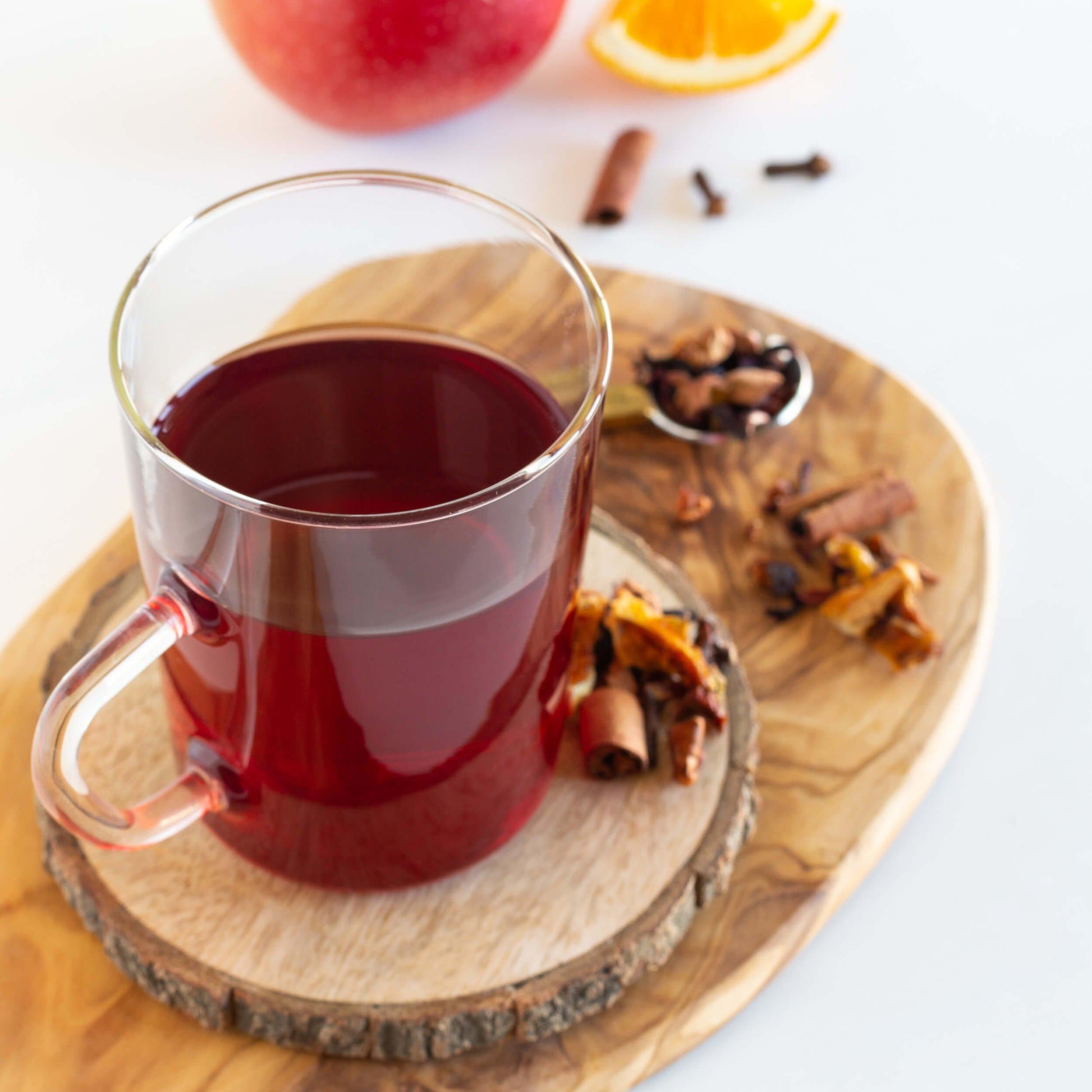 Mulled Wine Fruit Blend Herbal Tea shown as brewed tea in a glass mug, displayed on a wooden coaster with cinnamon and other ingredients nearby