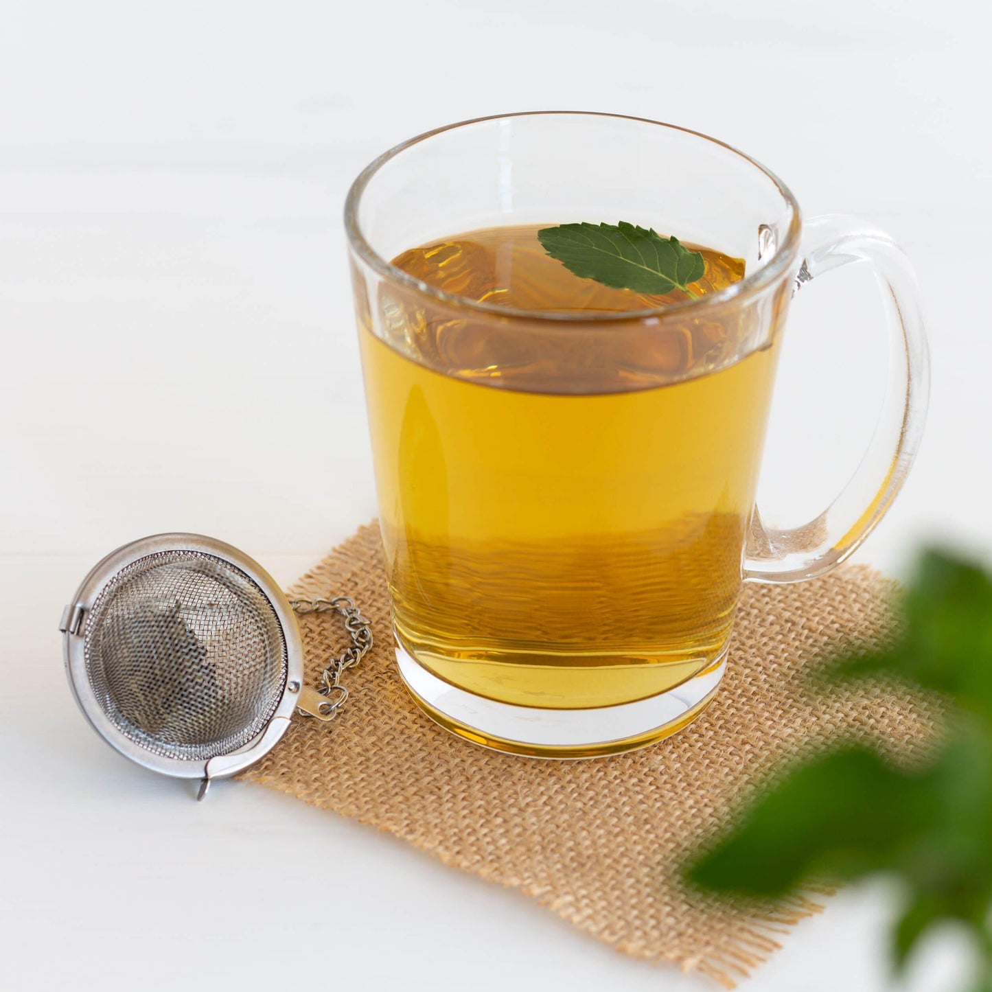 Peppermint Organic Herbal Tea shown as brewed tea in a glass mug, displayed on a square of burlap with a mesh teaball and mint leaves