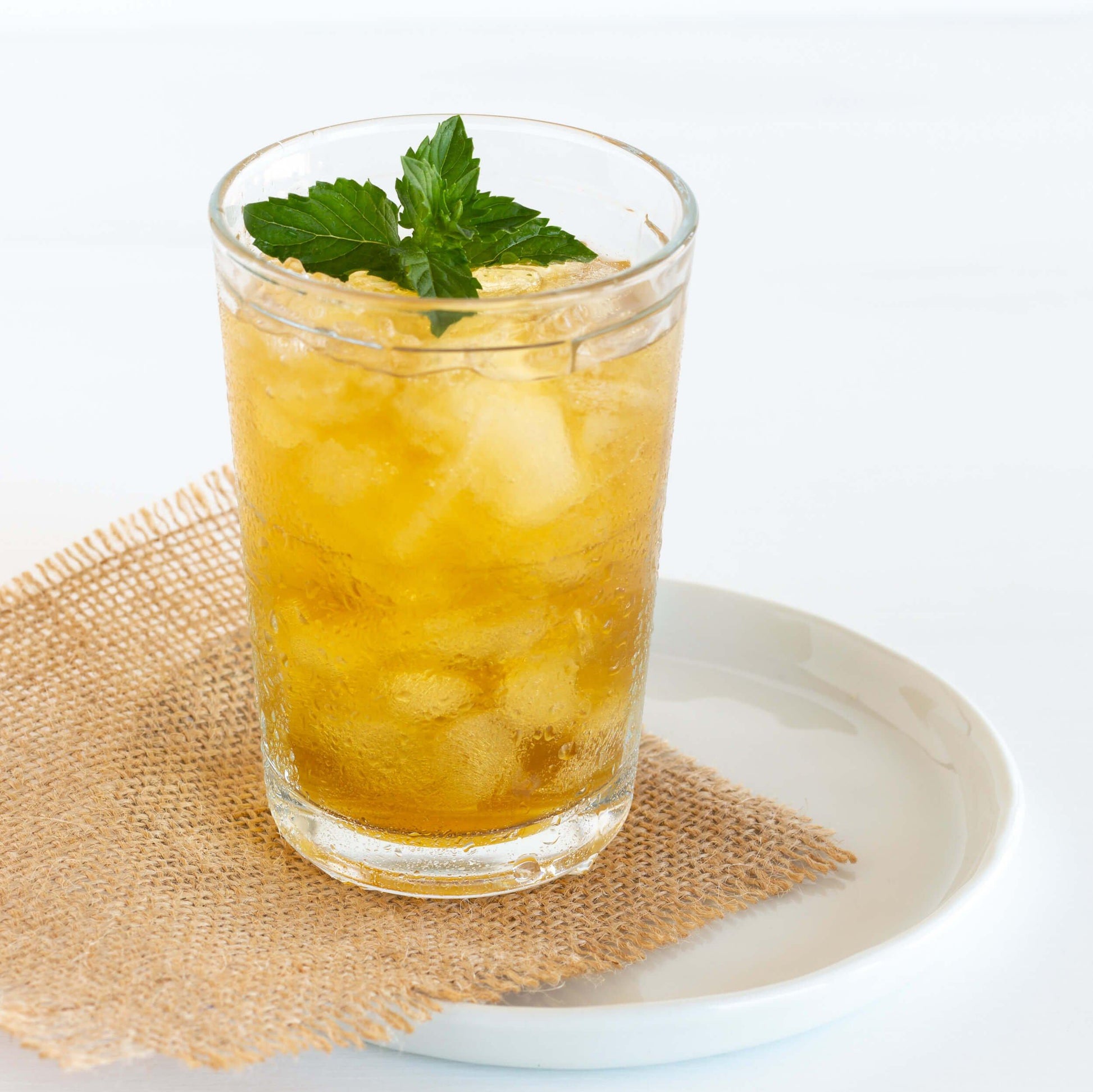 Peppermint Organic Herbal Tea shown as iced tea in a clear glass with a sprig of mint leaves, displayed on a square of burlap on a white saucer