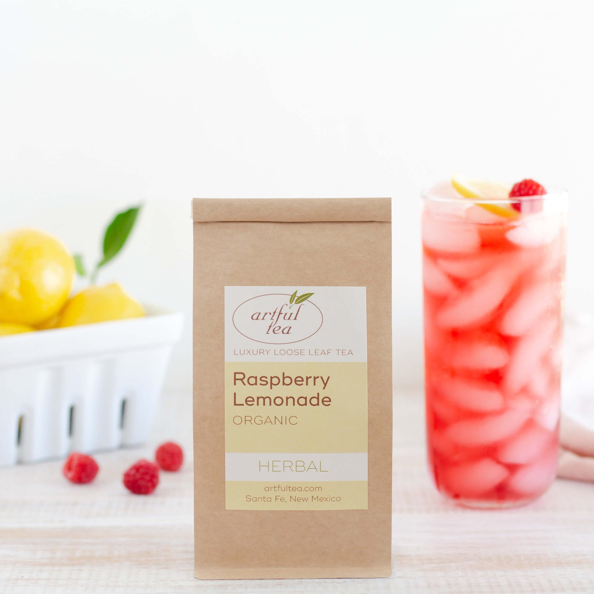 Raspberry Lemonade Organic Herbal Tea shown packaged in a kraft bag. A tall glass of iced tea and a container of lemons are in the background, with three raspberries scattered nearby