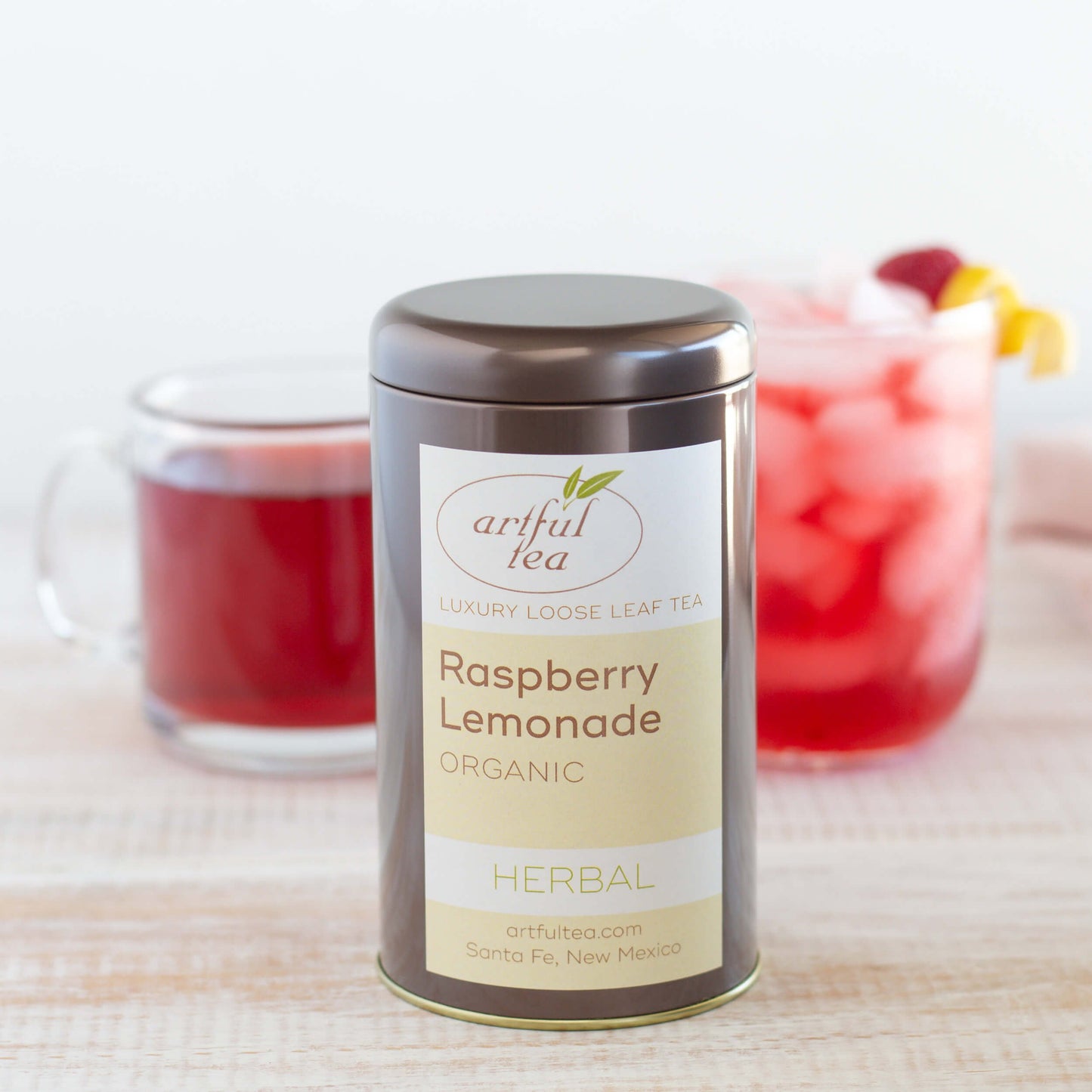 Raspberry Lemonade Organic Herbal Tea shown packaged in a brown tin, with a glass mug of brewed tea and a short glass of iced tea in the background