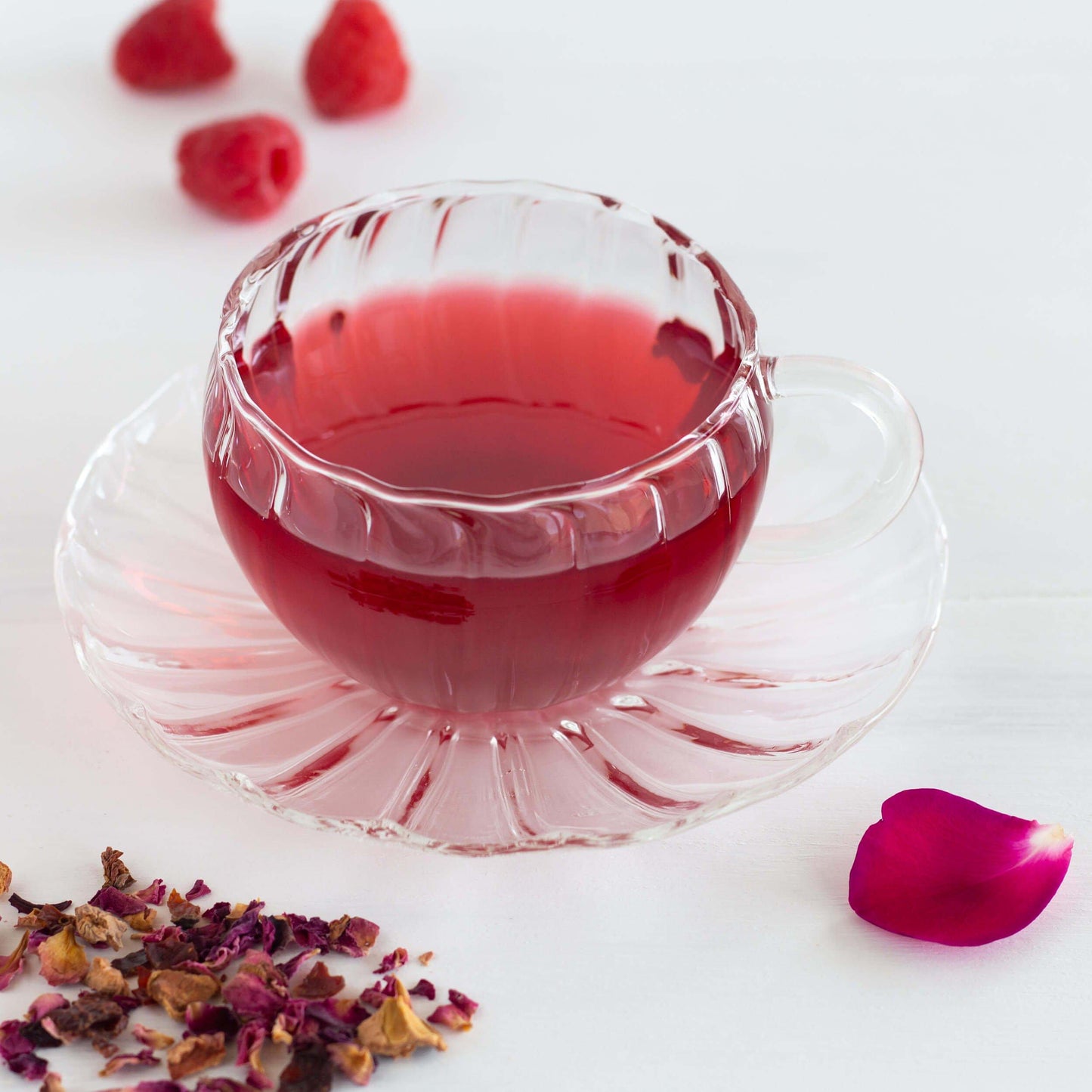 Rose Petal Raspberry Herbal Tea shown as brewed tea in a clear glass teacup and saucer, with loose tea leaves and a single red rose petal in the foreground, and three raspberries in the background