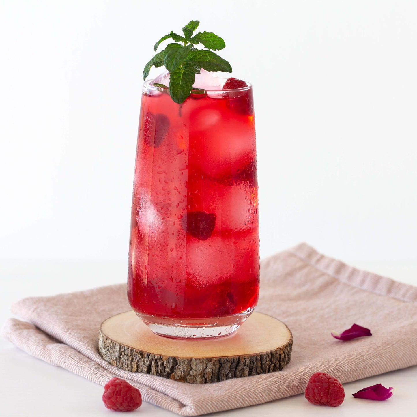 Rose Petal Raspberry Herbal Tea shown as iced tea in a clear glass with a sprig of mint and some raspberries floating in the glass. It is displayed on a wooden coaster on top of a piece of linen fabric