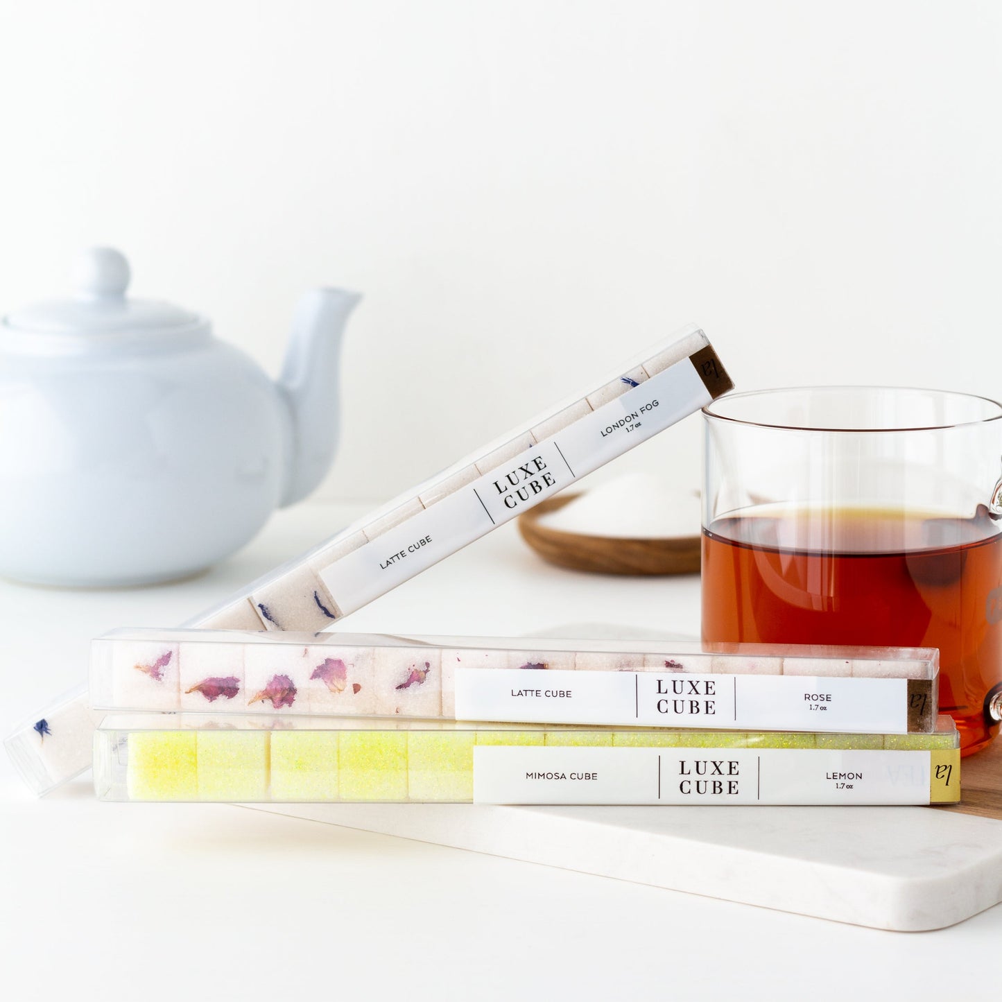 Luxe Sugar Cubes. Packages of 3 flavors (London Fog, Rose, and Lemon) displayed near a mug of tea, with white teapot in background.
