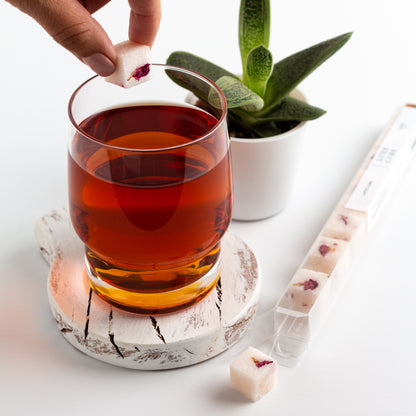 Luxe Sugar Cubes. A hand dropping one Rose sugar cube into a glass of tea, with an open package of Rose flavor sugar cubes nearby..