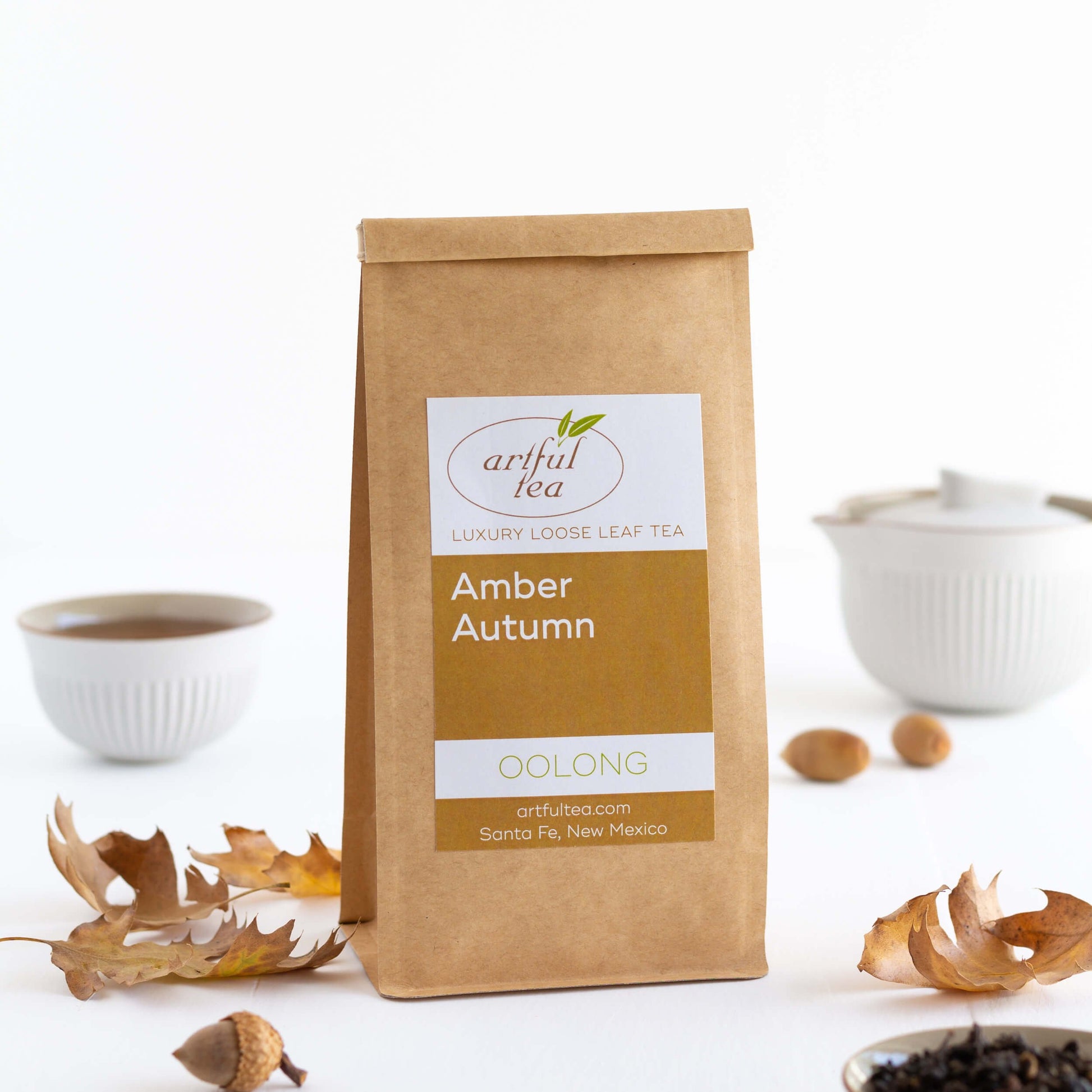 Amber Autumn Oolong shown packaged in a kraft bag, with acorns and brown tree leaves nearby, and a tiny white teacup and teapot in the background.