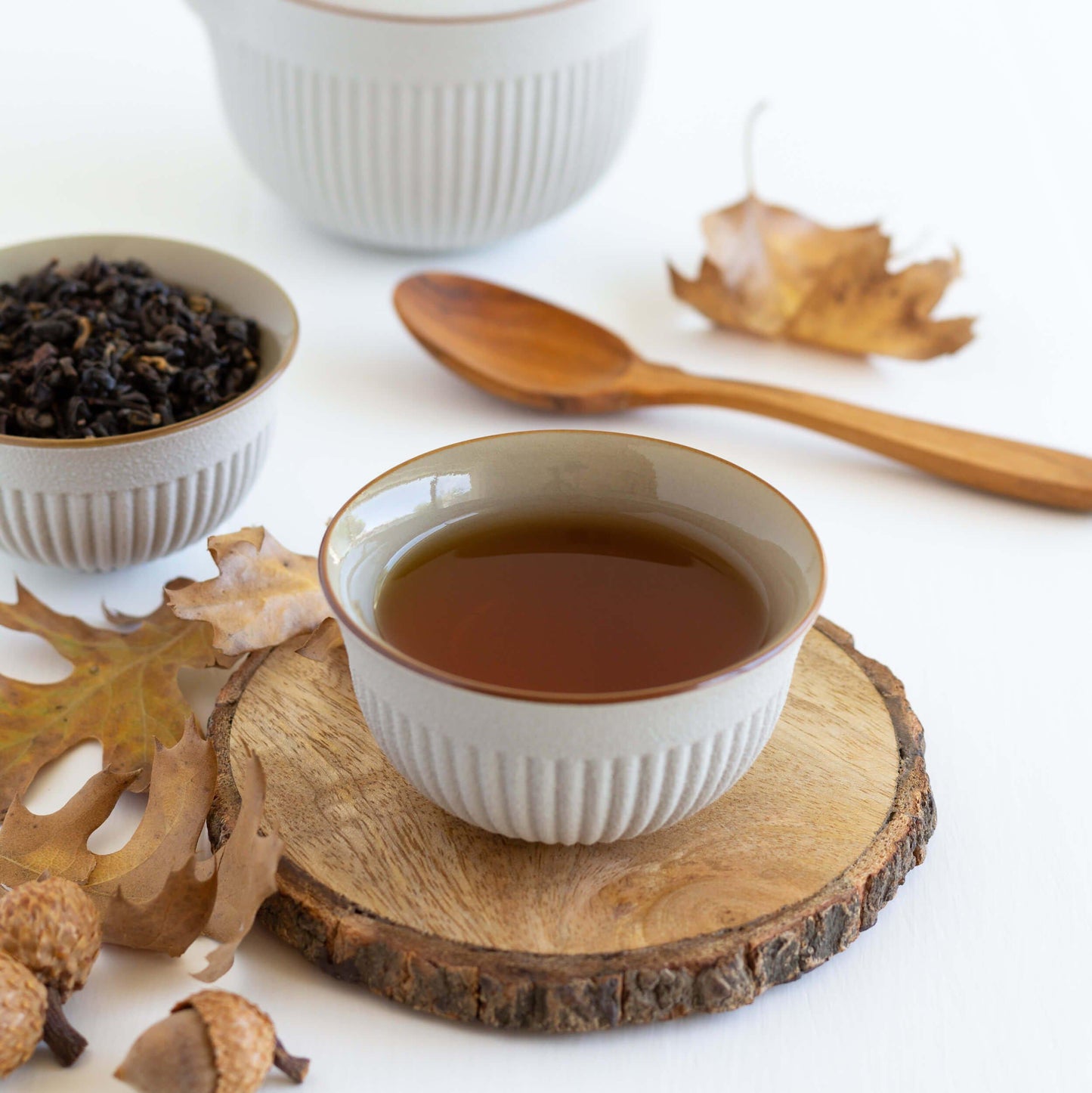Amber Autumn Oolong shown as brewed tea in a small white cup on a wooden coaster, with acorns, brown tree leaves, and a wooden spoon nearby.