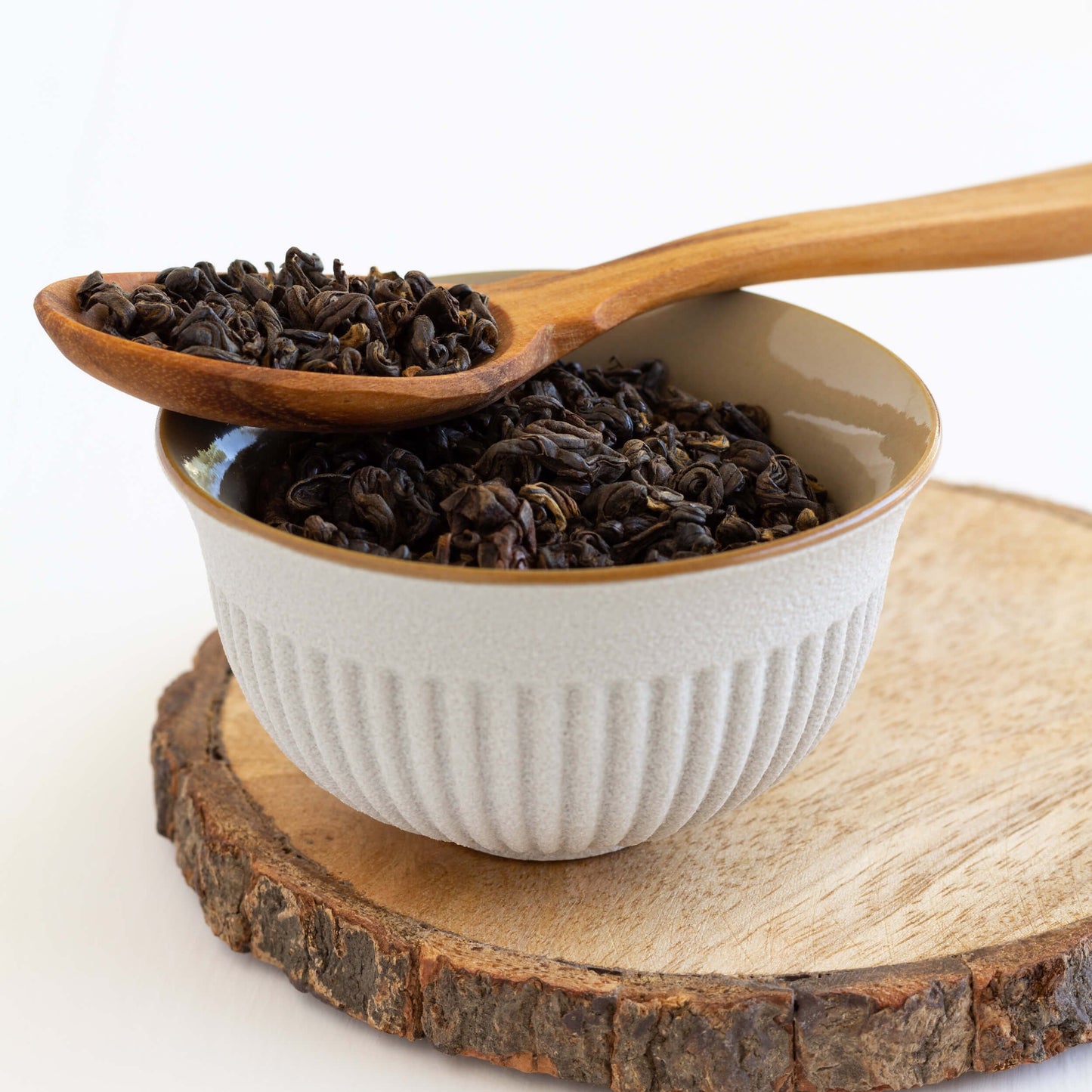Amber Autumn Oolong shown in close up as loose tea leaves in a wooden spoon, perched across a small white cup full of tea leaves, displayed on a wooden coaster.