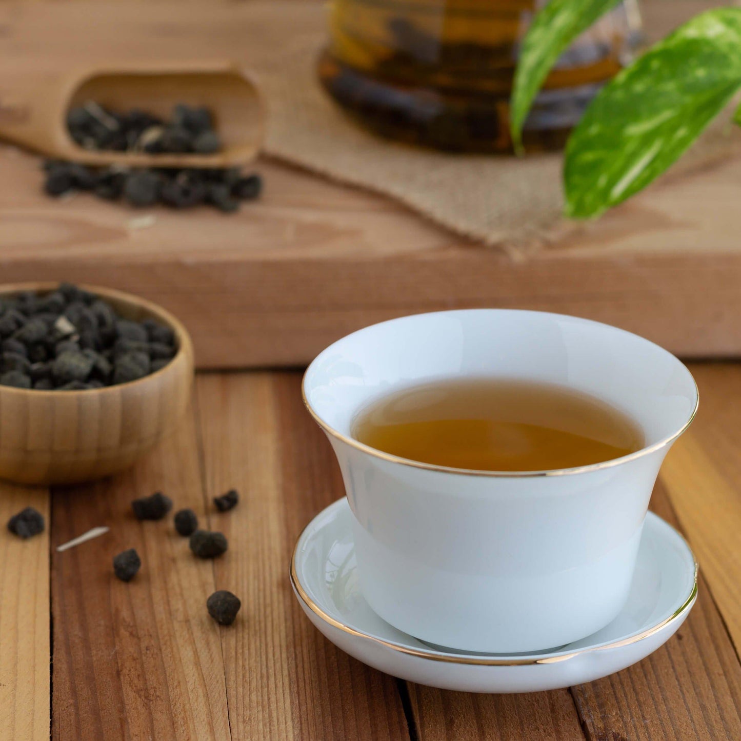 Ginseng Oolong Tea shown as brewed tea in a white teacup with gold rim. Loose tea leaves in the form of tiny rolled balls are displayed in the background