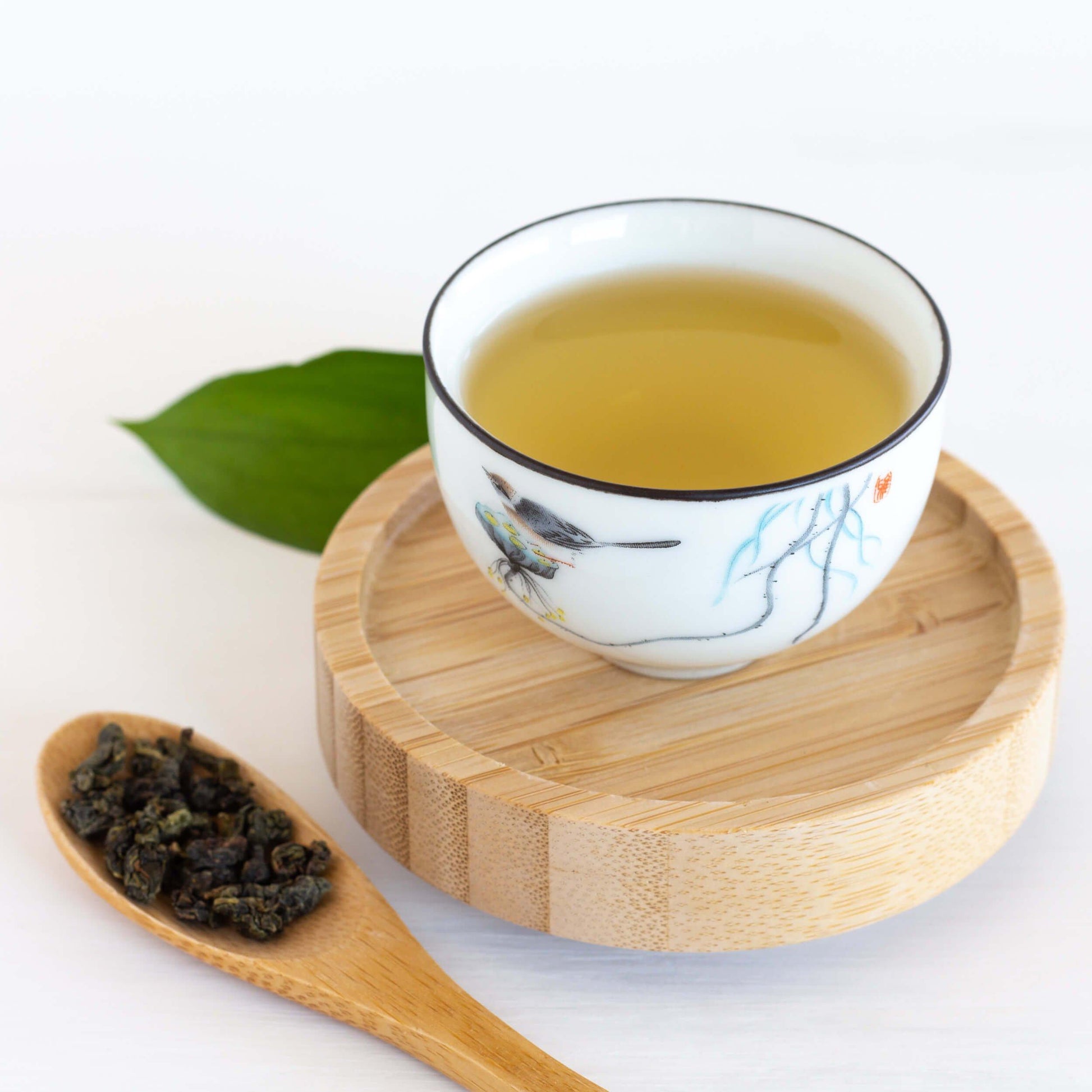 Jade Song Oolong Tea shown as brewed tea in a tiny white teacup with a bird design, displayed on a round bamboo coaster with a spoon of loose tea leaves