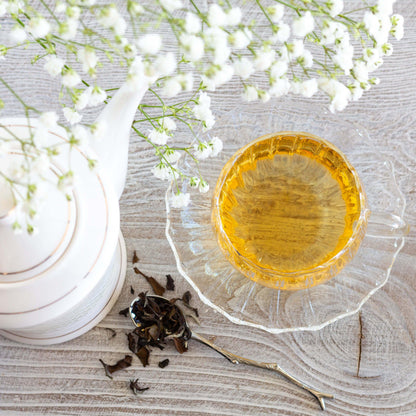 Oriental Beauty Oolong Tea shown from above as brewed tea in a clear glass teacup and saucer. A twig-shaped spoon of loose tea leaves and a whitte teapot are nearby. Baby's breath flowers are positioned above the scene