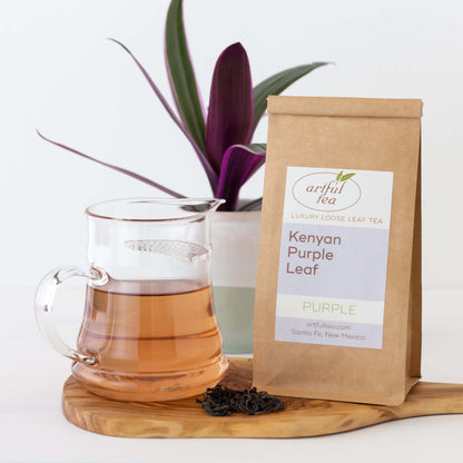 Kenyan Purple Leaf Tea shown packaged in a kraft bag next to a glass pitcher of brewed tea, displayed on a wooden board with loose tea leaves. A green and purple plant is in the background