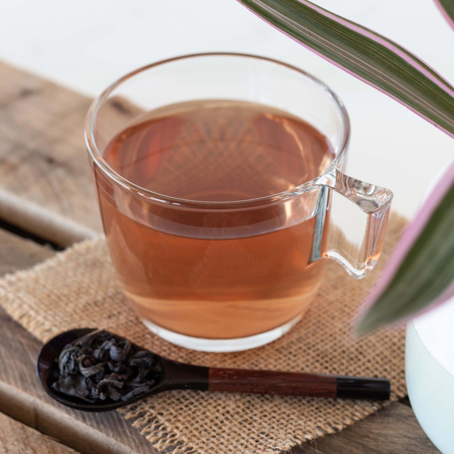 Kenyan Purple Leaf Tea shown as brewed tea in a glass mug with a spoonful of loose tea leaves in the foreground, all displayed on a square of burlap on wood slats