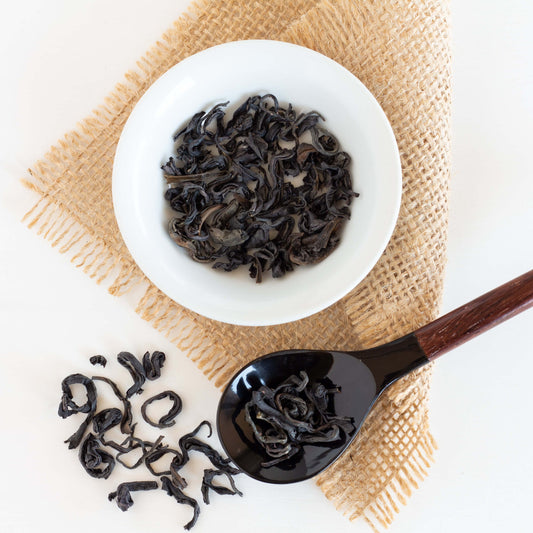 Kenyan Purple Leaf Tea shown from above a loose tea leaves in a white dish and in a black spoon, both displayed on a piece of burlap