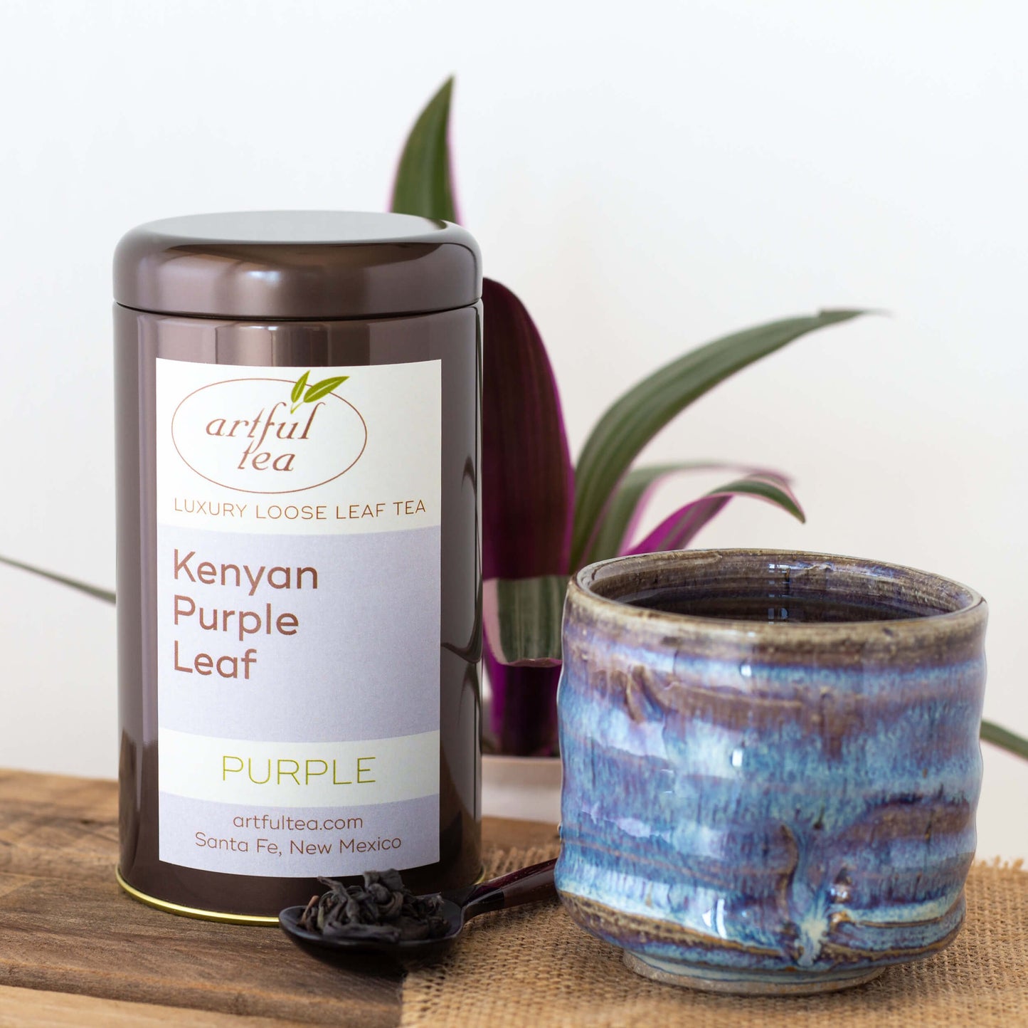 Kenyan Purple Leaf Tea shown packaged in a brown tin next to a blue pottery teacup and a spoon of tea leaves