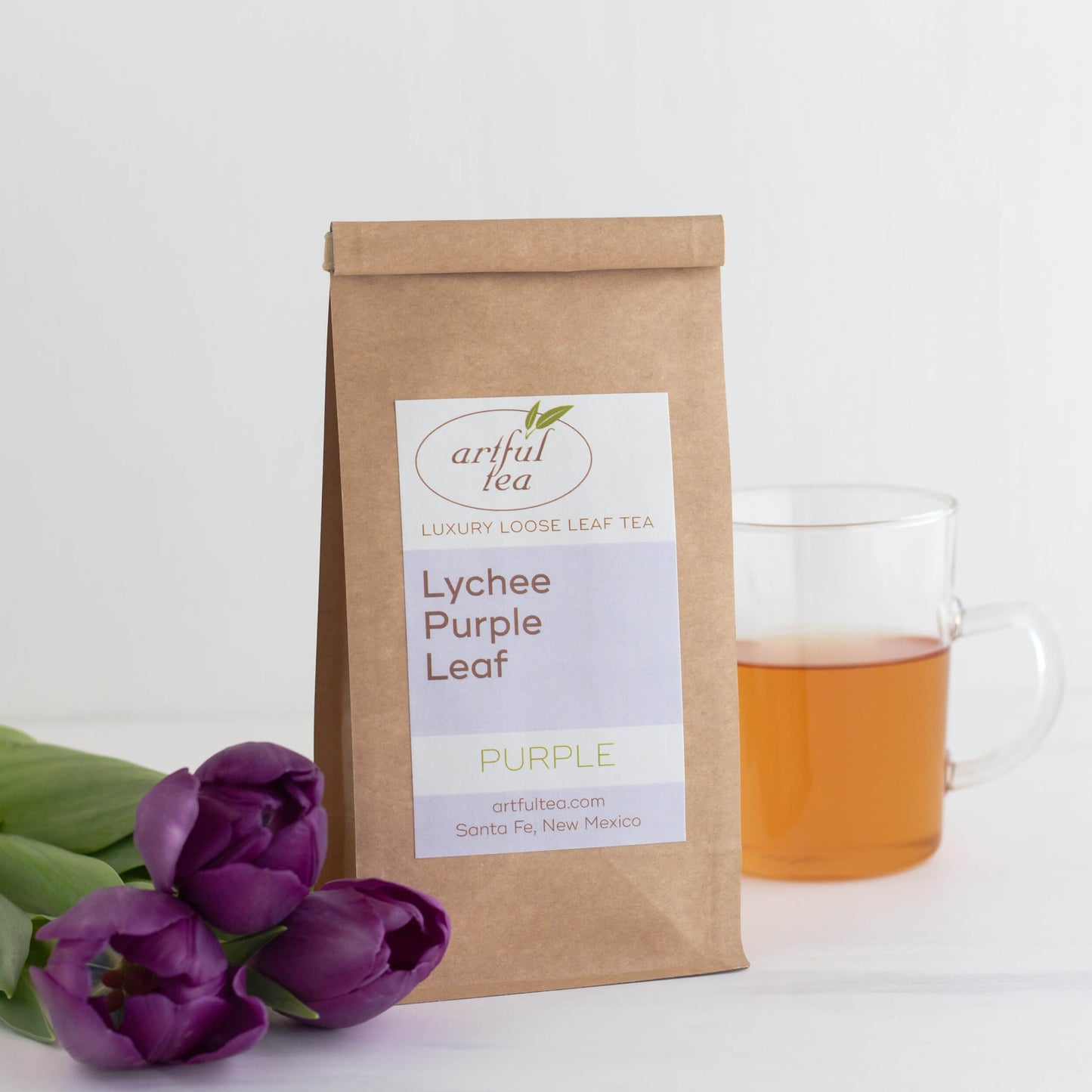 Lychee Purple Leaf tea shown packaged in a kraft bag, with three purple tulips on the left and a glass mug of brewed tea on the right