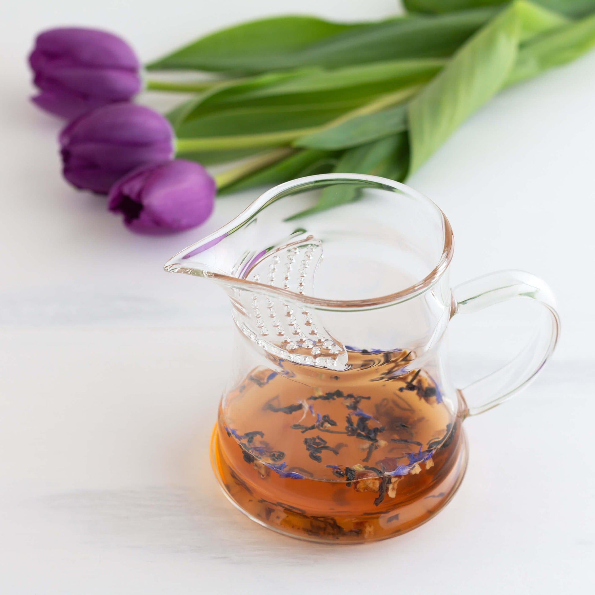 Lychee Purple Leaf Tea shown brewed in a glass pitcher with three purple tulips in background