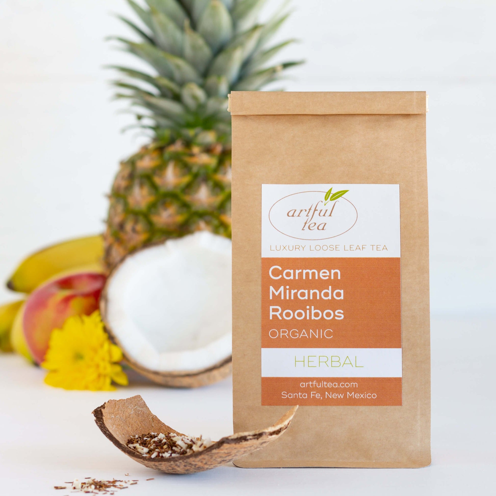 Carmen Miranda Rooibos Organic Herbal Tea shown packaged in a kraft bag, with a piece of coconut shell and loose tea in the foreground, and a coconut half, a pineapple, an apple, and bananas in the background