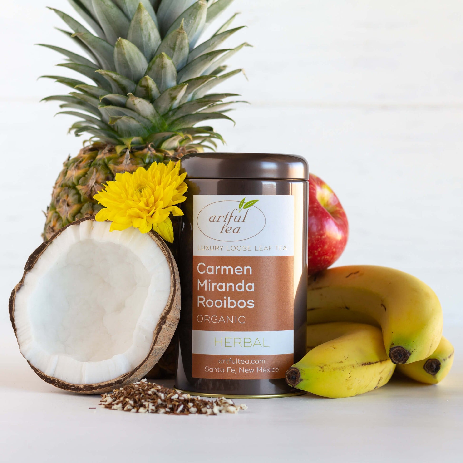 Carmen Miranda Rooibos Organic Herbal Tea shown packaged in a brown tin, surrounded by bananas, a coconut half, a pineapple, an apple, and a yellow flower