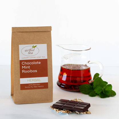 Chocolate Mint Rooibos herbal tea shown packaged in a kraft bag, with a bar of chocolate in the foreground and a pitcher of brewed tea with mint leaves in the background
