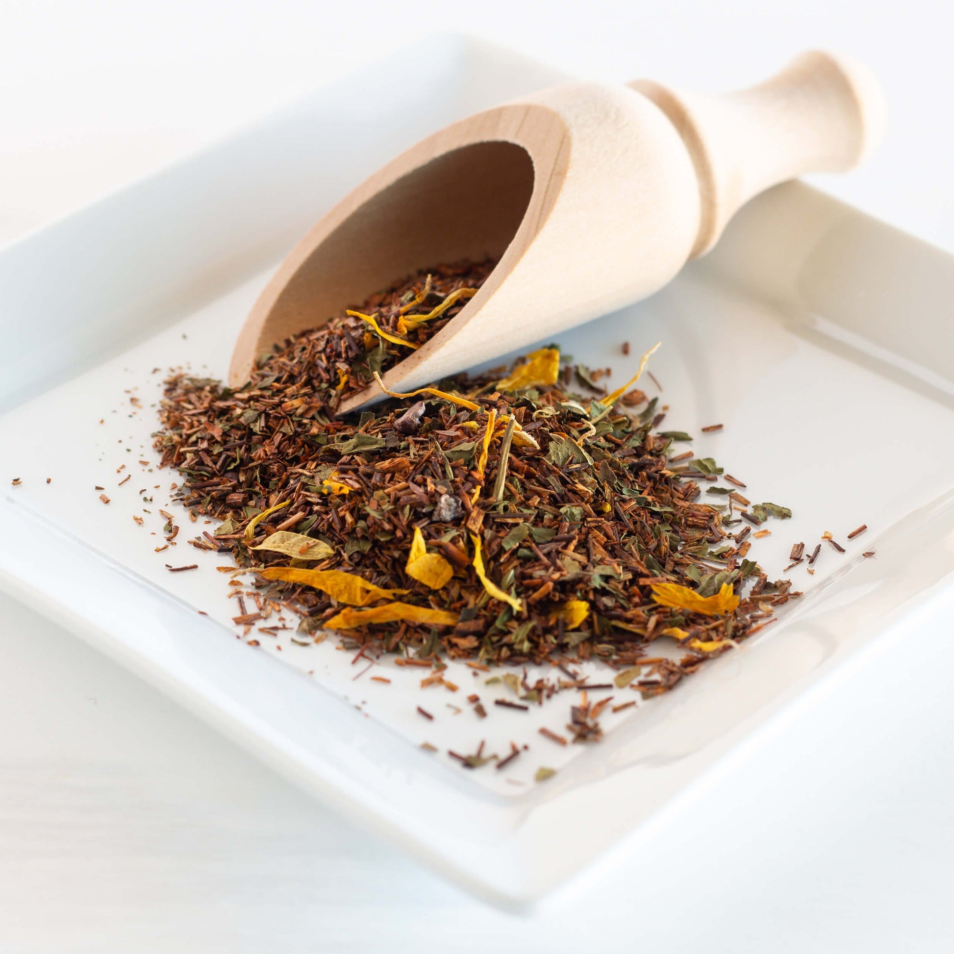 Chocolate Mint Rooibos herbal tea shown as loose tea leaves spilling from a wooden scoop on a square white plate