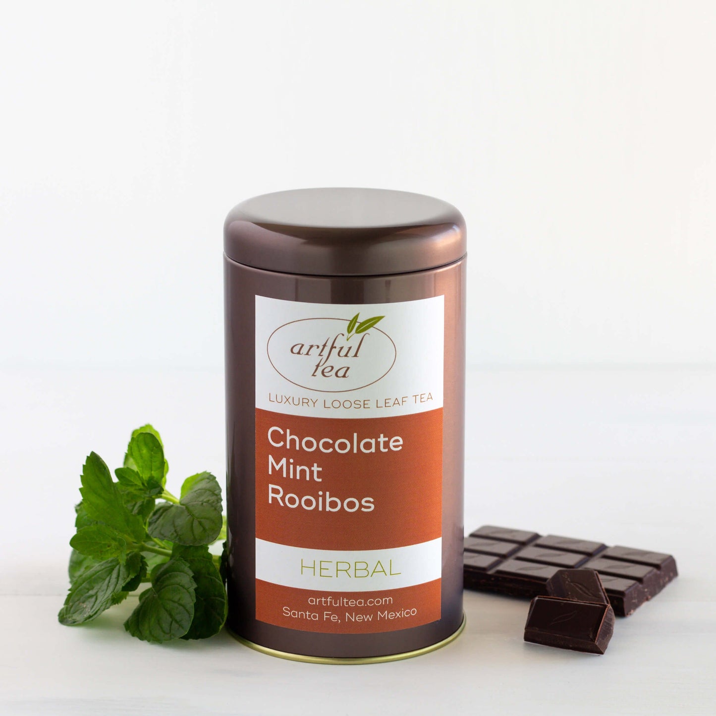 Chocolate Mint Rooibos herbal tea shown packaged in a brown tin, surrounded by fresh mint leaves and a bar of chocolate