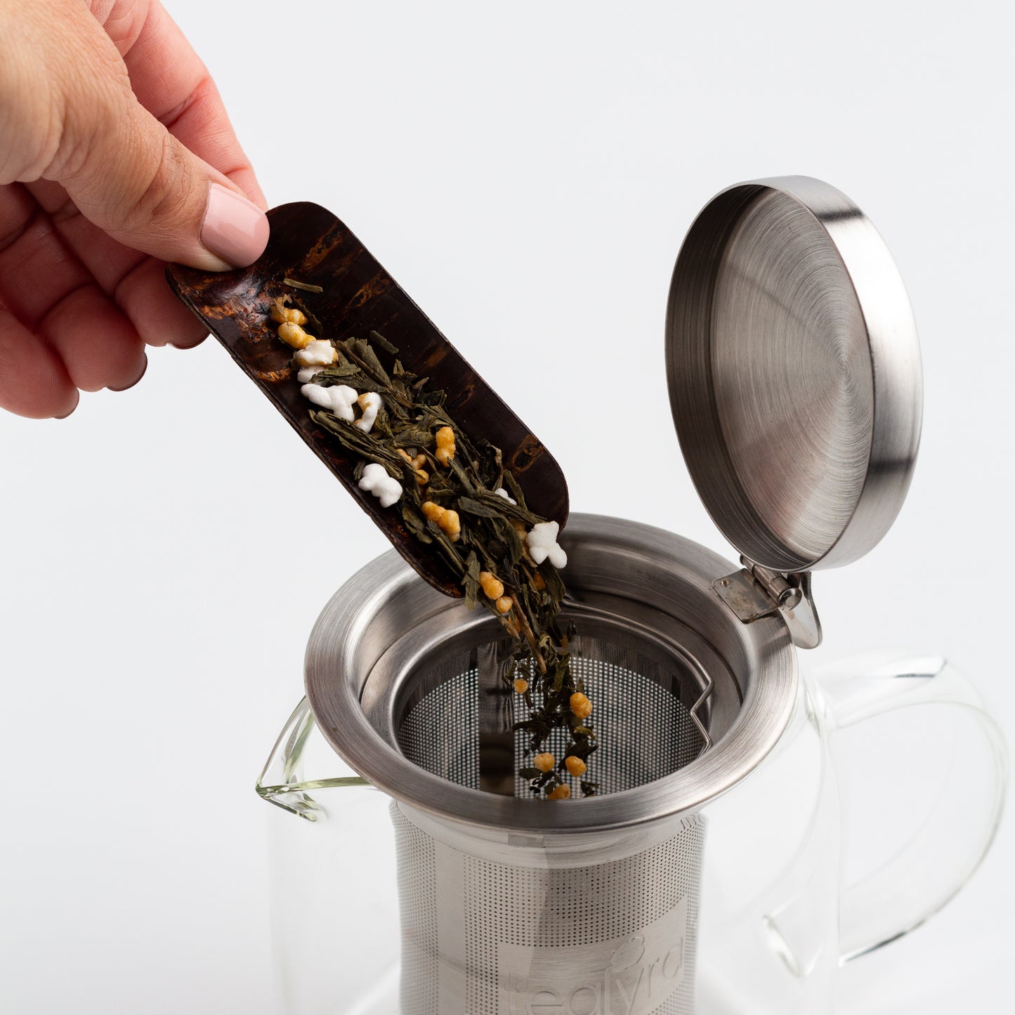 A hand shown using a cherry wood tea scoop to pour tea leaves into a stainless steel infuser in a glass teapot.