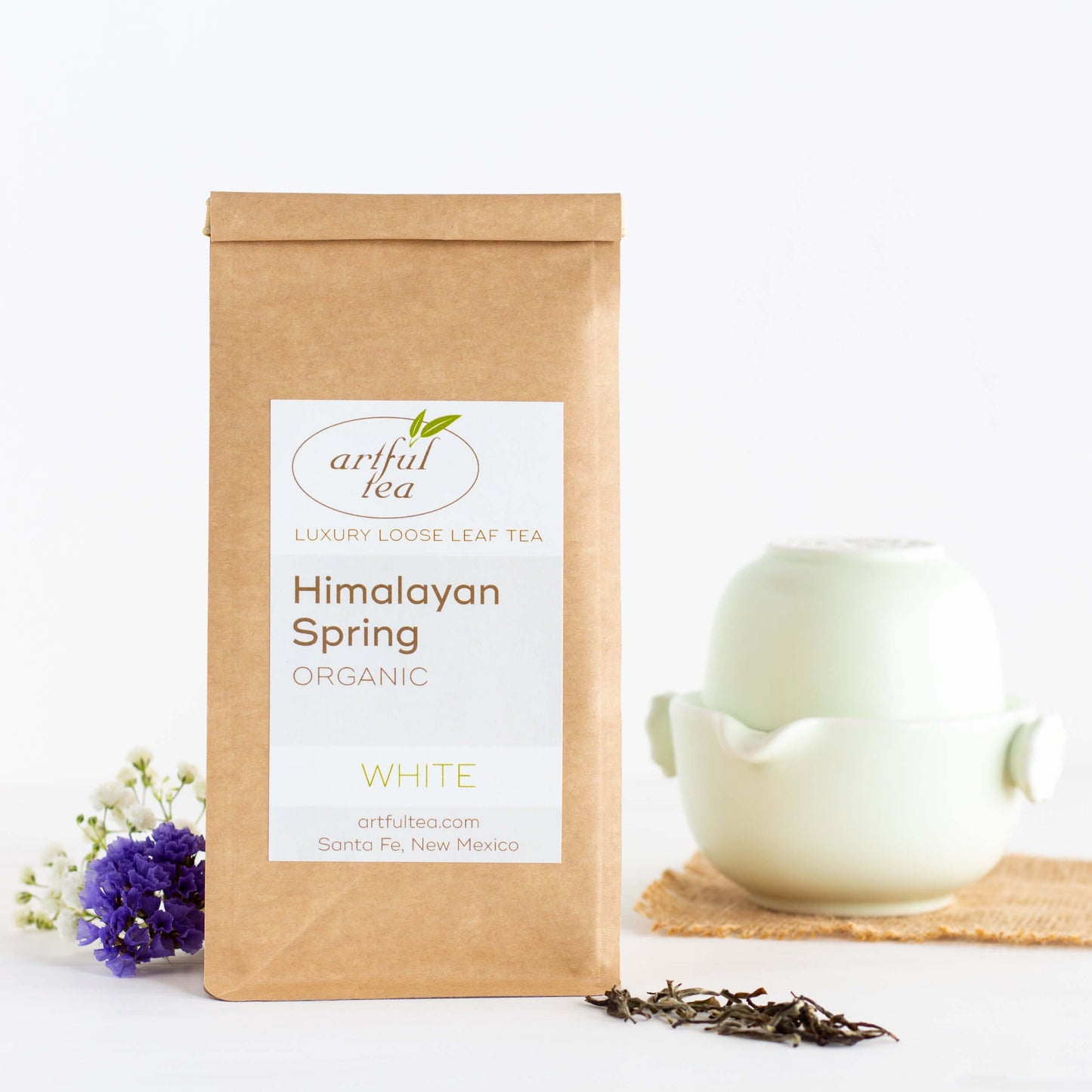 Himalayan Spring Organic White Tea shown packaged in a kraft bag, with a small pale green teapot and cup set in the background, and loose tea leaves in the foreground.