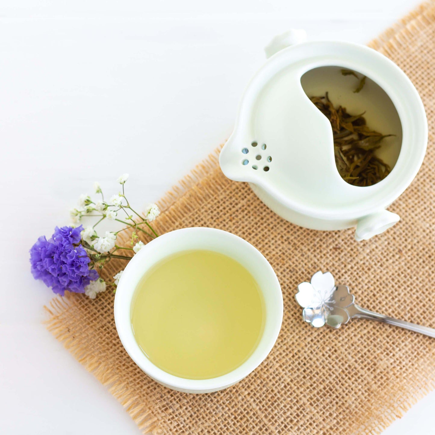 Himalayan Spring Organic White Tea shown from above brewing in a small pale green teapot and teacup with a flower-shaped spoon displayed on burlap.