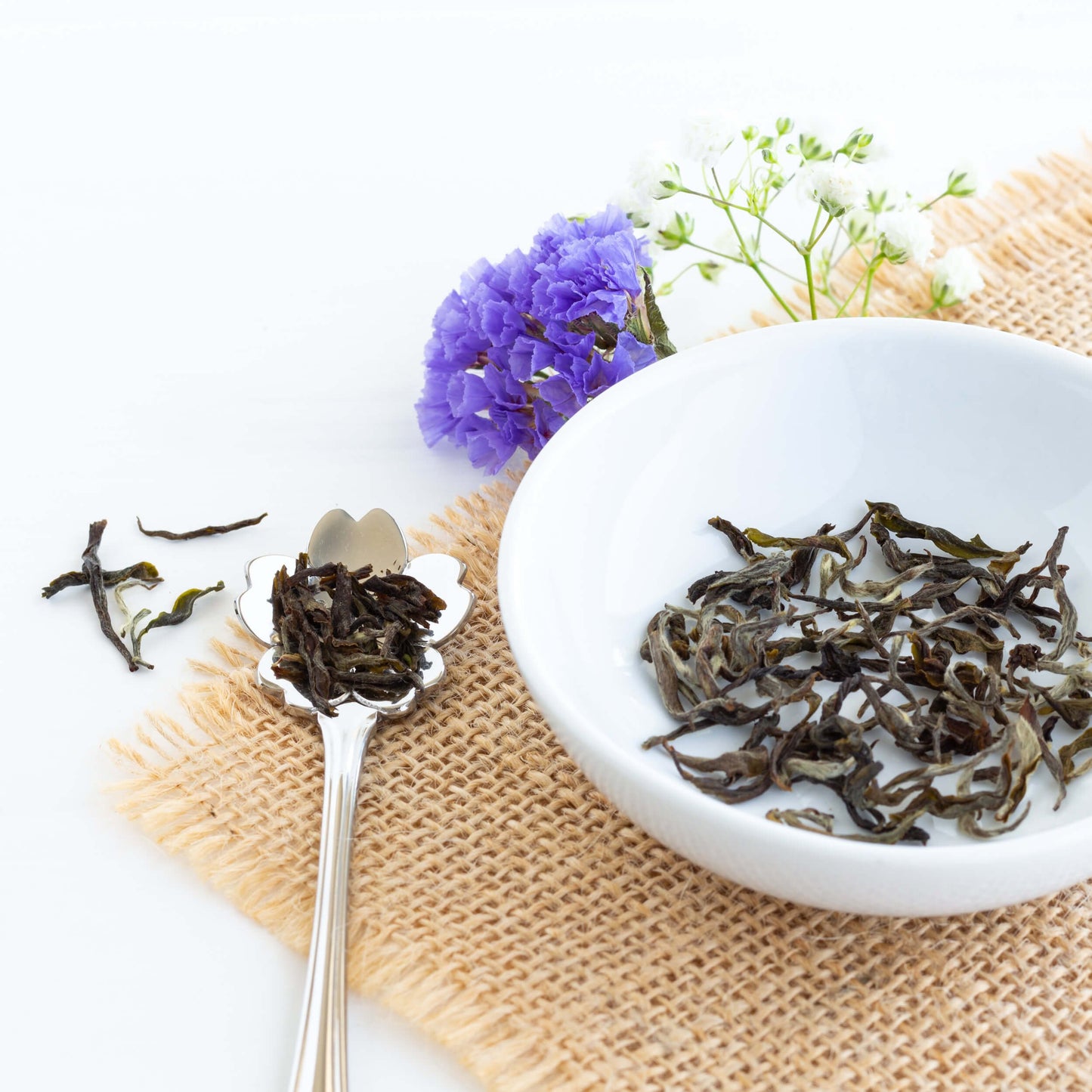 Himalayan Spring Organic Green Tea shown as loose tea leaves in a white dish with a flower-shaped spoon nearby, displayed on burlap with some purple flowers.