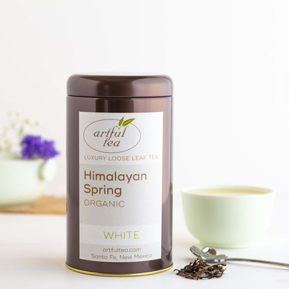 Himalayan Spring Organic White Tea shown packaged in a brown tin, with loose tea leaves nearby. A pale green teacup and some small flowers are in the background.