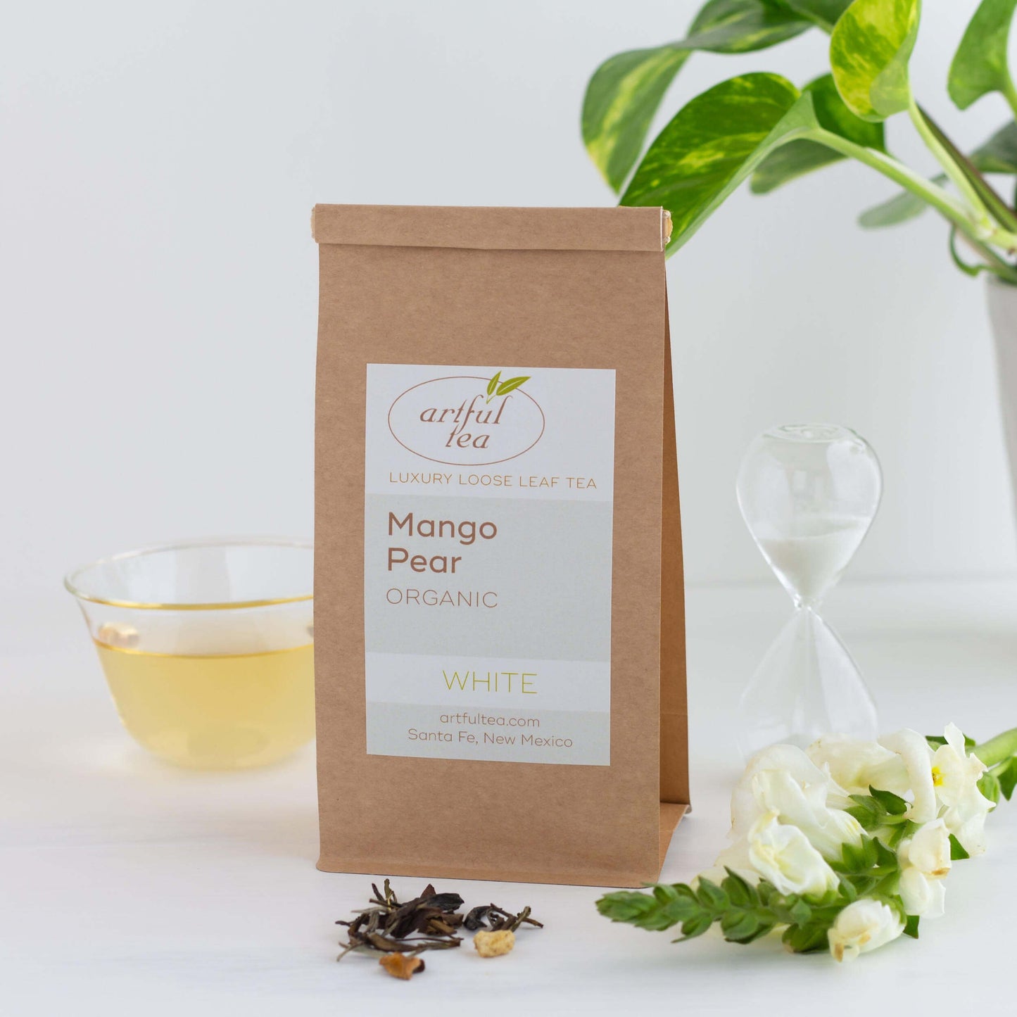 Mango Pear Organic White Tea shown packaged in a kraft bag with some white flowers and loose tea leaves in the foreground. A small sand timer, a green plant, and a small glass of brewed tea are in the background