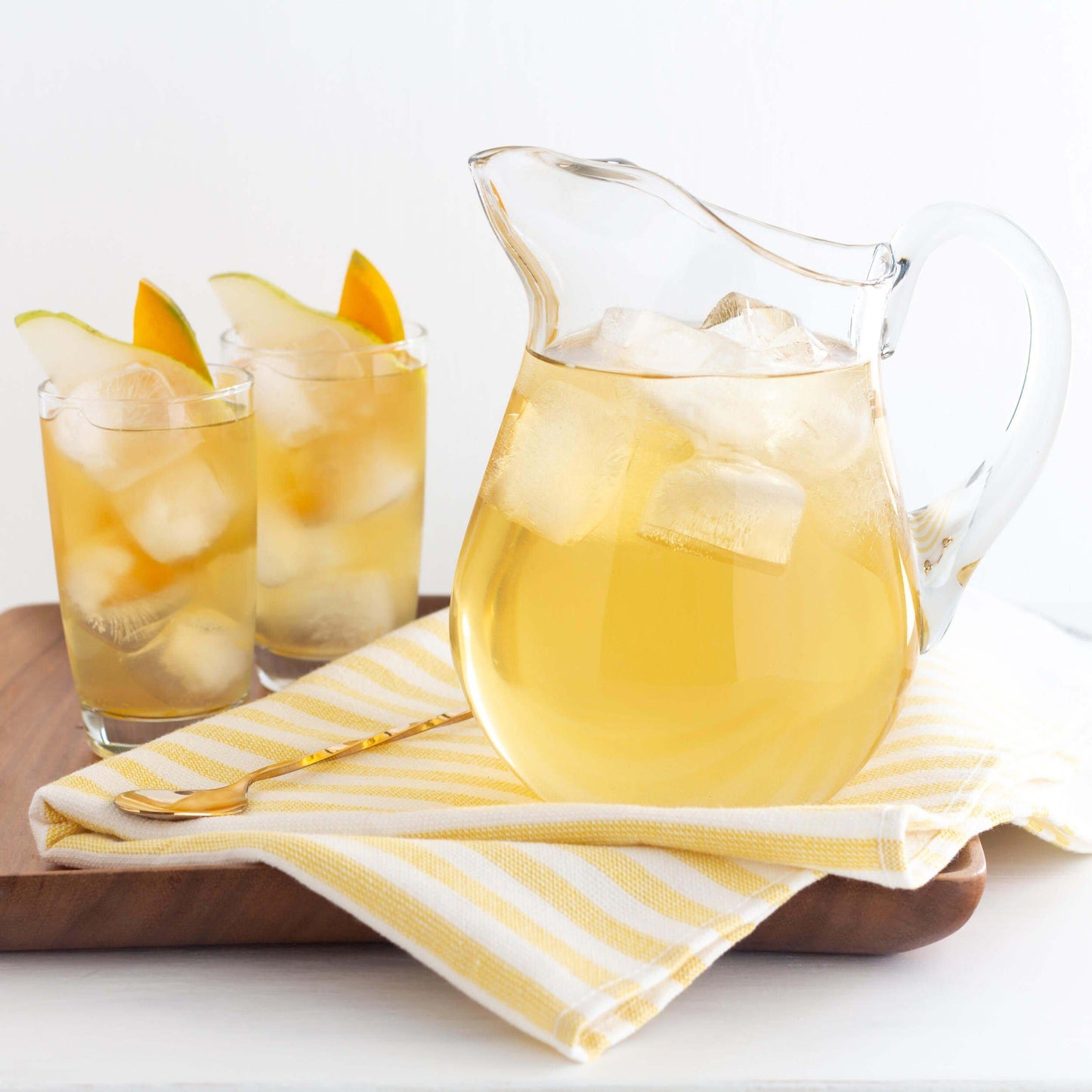 Mango Pear Organic White Tea shown as iced tea in a glass pitcher, with two glasses of iced tea garnished with mango wedges, displayed on yellow and white striped towel