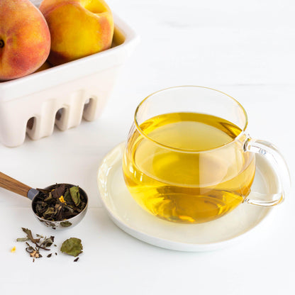 Peachy Keen Organic White Tea shown as brewed tea in a glass mug on a white saucer, with a scoop of loose tea leaves and a small basket of peaches