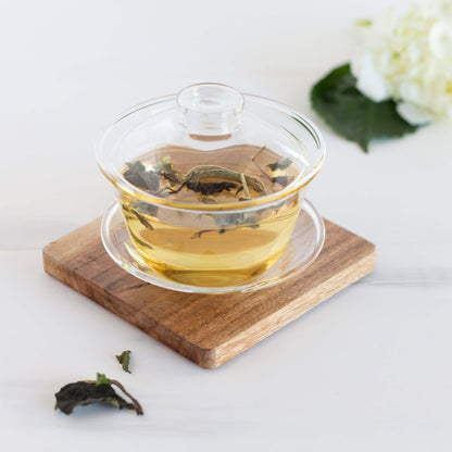 White Peony Organic White Tea shown as brewed tea in a glass gaiwan cup with a lid, displayed on a wooden coaster